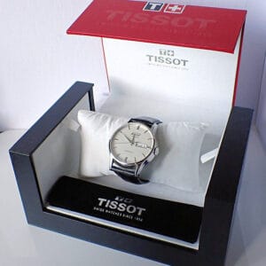 A Tissot Visodate Automatic 2012 wristwatch with a silver face and black leather strap is displayed on a white cushion inside an open Tissot box showing the brand name and logo.