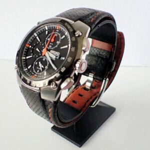The Seiko Sportura Alarm Chronograph 100m, featuring a black dial, perforated leather strap, silver case, and multiple dials, is displayed upright on a black stand against a white background.