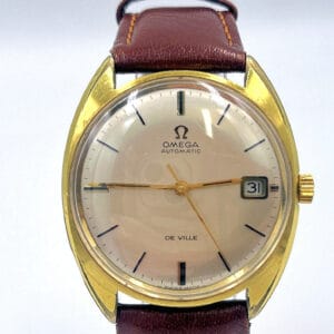 The Omega De Ville Automatic Date 1970 is a gold wristwatch with a brown leather strap, white dial, gold hands, black hour markers, date window at “31,” and features the inscriptions OMEGA AUTOMATIC and DE VILLE on the face.