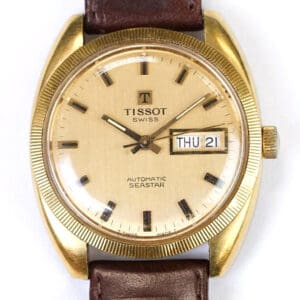 The Tissot Seastar Automatic Gold Plated Day Date Watch features a gold-toned round case, beige dial with black hour markers, day and date window (THU 21), and a brown leather strap.