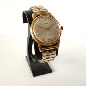 The Ilford Pie Pan Mechanical 1960-1969 wristwatch features a gold-tone case, silver dial, and a gold and silver metal strap, displayed upright on a black stand against a plain white background.