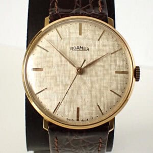 A Roamer Mechanical 1970s Linen Dial wristwatch with a textured gold-toned round dial, slim gold markers, and a brown leather strap, displayed upright on a black stand against a plain background.