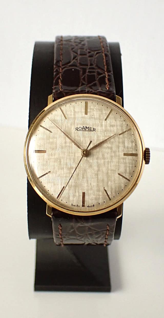 A Roamer Mechanical 1970s Linen Dial wristwatch with a textured gold-toned round dial, slim gold markers, and a brown leather strap, displayed upright on a black stand against a plain background.