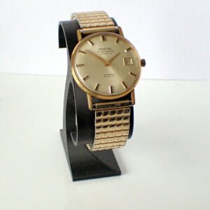 A Montine Automatic Date 1970s gold wristwatch with a square-patterned metal strap, round face, and date display is shown upright on a black stand against a white background.
