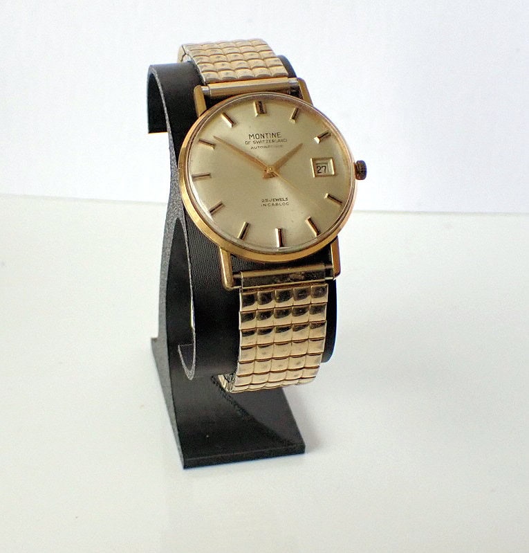 A Montine Automatic Date 1970s gold wristwatch with a square-patterned metal strap, round face, and date display is shown upright on a black stand against a white background.