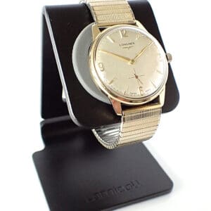 A Longines 9ct Gold Calibre 370 Presentation Watch (1963) is shown upright on a black Lamicall stand against a white background. The watch features a round gold-tone face, simple markers, and a small seconds sub-dial.