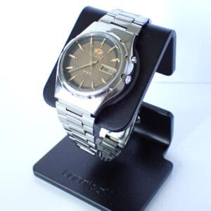The Orient Automatic Day Date is a silver analogue wristwatch with a metal strap, gold hour markers, day and date windows, and brand logo on the dial. Displayed on a black stand against a white background.