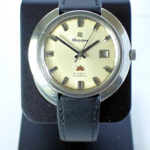 A Ricoh 'Space Age' Sakura Automatic Date 1970s watch with a round silver case, cream dial marked "21 Jewels Automatic," date window, and black leather strap is displayed on a black stand.