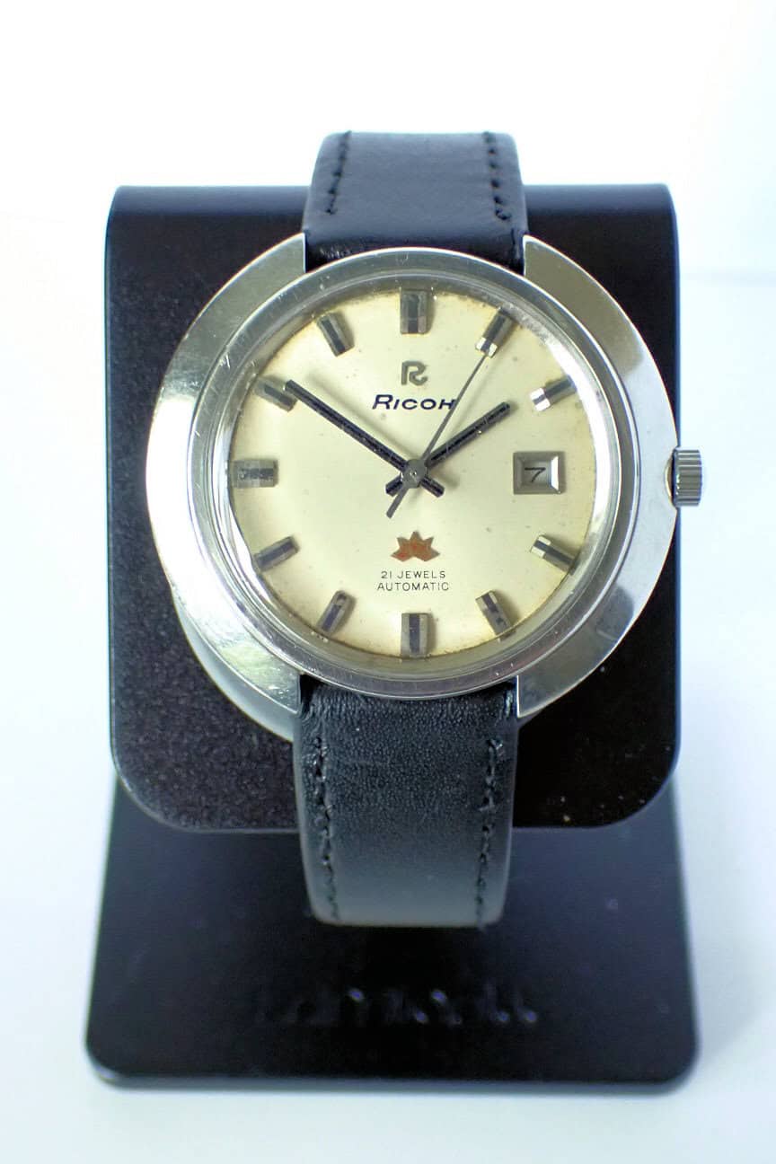 A Ricoh 'Space Age' Sakura Automatic Date 1970s watch with a round silver case, cream dial marked "21 Jewels Automatic," date window, and black leather strap is displayed on a black stand.
