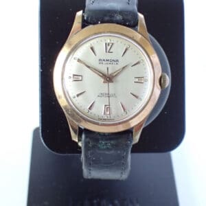 A Ramona Early 1958 Automatic 25 Jewels Watch with a gold-tone round case, silver dial, black leather strap, and date window, displayed upright on a black watch stand.