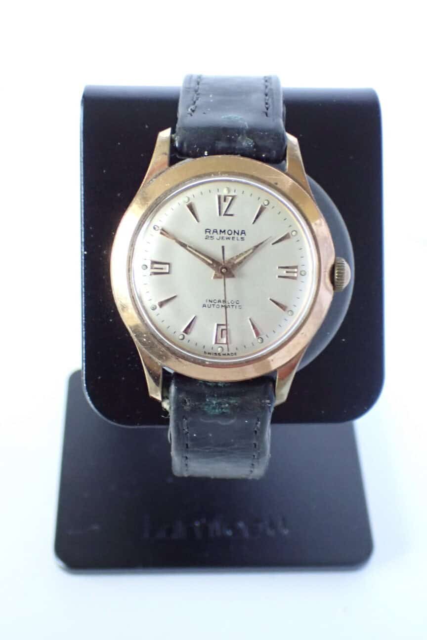 A Ramona Early 1958 Automatic 25 Jewels Watch with a gold-tone round case, silver dial, black leather strap, and date window, displayed upright on a black watch stand.
