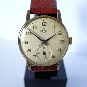 A Smiths De Luxe Mechanical 9ct Gold 1950s wristwatch featuring a gold case, cream dial with Arabic numerals, small seconds sub-dial, and textured brown leather strap is displayed on a stand.