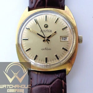 A Roamer Anfibio Mechanical Gold Plated 1960s wristwatch with a gold-tone round face, black hour markers, date window at 3 o’clock, brown leather strap, and ROAMER logo below 12 o’clock.