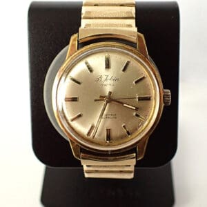 A vintage B JOBIN Gold Plated Mechanical Cal ST 96 1956 watch with a round face, gold hour markers, and metal strap is displayed on a black stand. The dial reads S. Jobin Swiss, noting 17 jewels and carbloc.