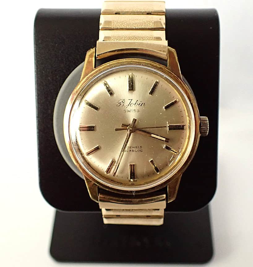 A vintage B JOBIN Gold Plated Mechanical Cal ST 96 1956 watch with a round face, gold hour markers, and metal strap is displayed on a black stand. The dial reads S. Jobin Swiss, noting 17 jewels and carbloc.