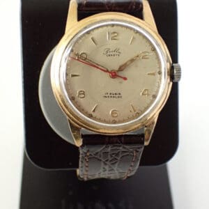 The Bella Genève Gold-Plated Mechanical 1950s wristwatch features a round gold-tone case, cream dial with simple hour markers, and a brown leather strap, displayed upright on a black stand.