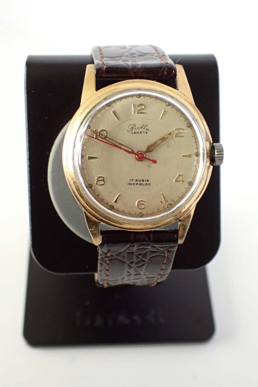 The Bella Genève Gold-Plated Mechanical 1950s wristwatch features a round gold-tone case, cream dial with simple hour markers, and a brown leather strap, displayed upright on a black stand.
