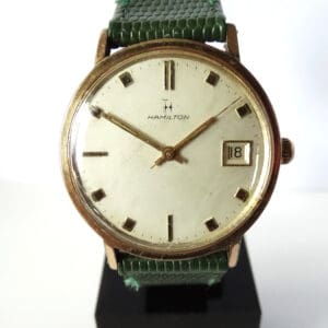 The Hamilton Date Mechanical Watch C1964 features a vintage round gold case, white dial with gold hour markers, a date window at 3 o'clock, green textured strap, and displays the time as 10:09.