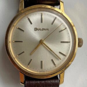 The Bulova Gold Plated Mechanical 1970s wristwatch features a gold-tone round case, silver sunburst dial with gold hour markers, and a brown leather strap, displaying the time as 4:33:53.