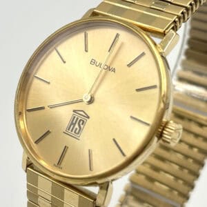 A gold Bulova Longchamp watch (Special Edition for H & S Investment Company in America) with a metallic strap, minimalist dial, black hour markers, no numbers, and an HS logo above a column at 6 o'clock. The hands show 10:07.