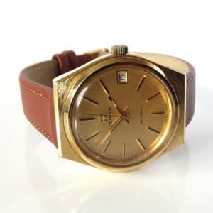 A Zenith Date Automatic Watch Calibre 2572 c1976 with a gold case, gold dial, brown leather strap, and date set to the 15th is shown on a white background.