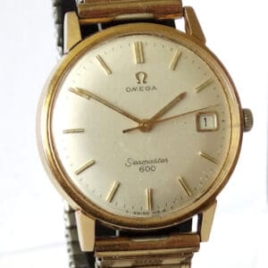 An Omega Seamaster 600 Date Manual Wind gold-plated watch with a round face, date display, and metal bracelet is displayed upright on a black stand. Pay interest-free in three with Klarna.
