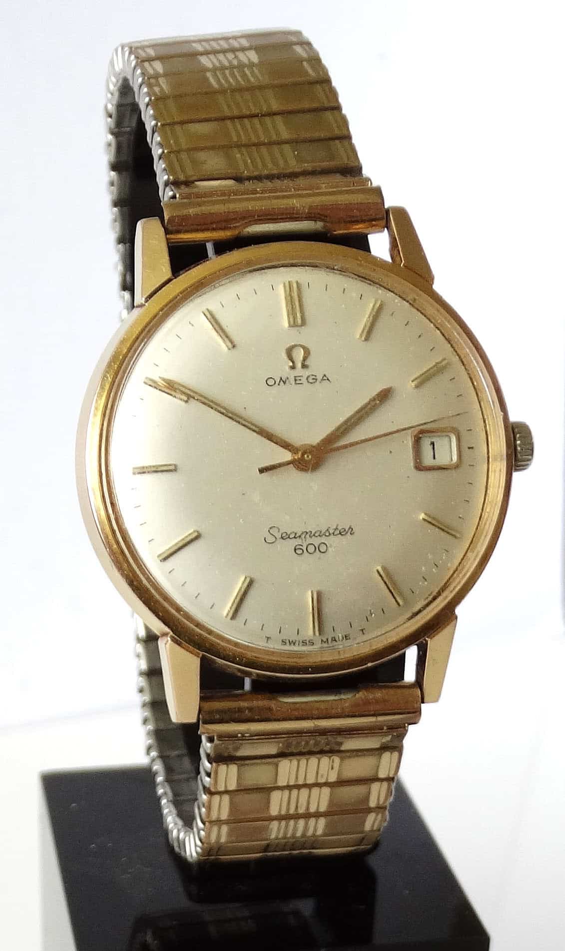 An Omega Seamaster 600 Date Manual Wind gold-plated watch with a round face, date display, and metal bracelet is displayed upright on a black stand. Pay interest-free in three with Klarna.