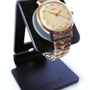 An Oris mechanical watch with a gold case, cream dial, gold Arabic numerals, and a gold expansion strap is displayed on a black stand.