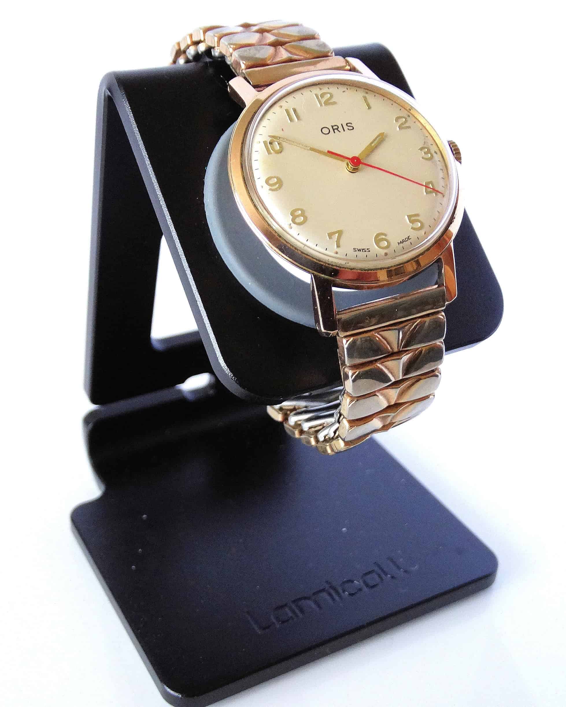 An Oris mechanical watch with a gold case, cream dial, gold Arabic numerals, and a gold expansion strap is displayed on a black stand.