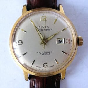 The Oris Date 654 KIF Watch features a vintage gold-tone case, white dial with ANTI-SHOCK 17 JEWELS text, date display, and a brown leather strap.