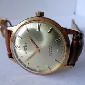 A Fortis 21 Jewels Incabloc - Quality Hand Wind 103150 2377 wristwatch with a gold-tone case, silver dial, brown leather strap, and bold numerals at 12, 3, 6, and 9 is displayed resting on a white surface.