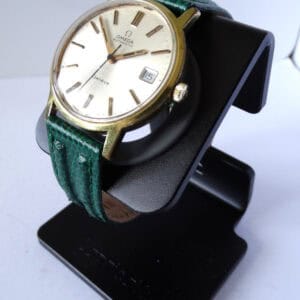 An Omega Genève Gold Tone Date Watch Cal 1012 with a white dial and green textured leather strap, shown on a black stand. Pay interest-free in three with Klarna.