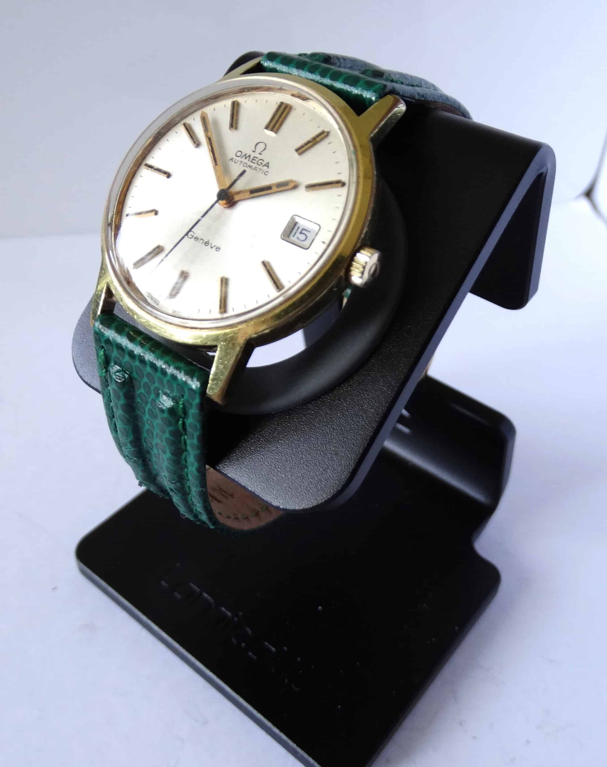 An Omega Genève Gold Tone Date Watch Cal 1012 with a white dial and green textured leather strap, shown on a black stand. Pay interest-free in three with Klarna.