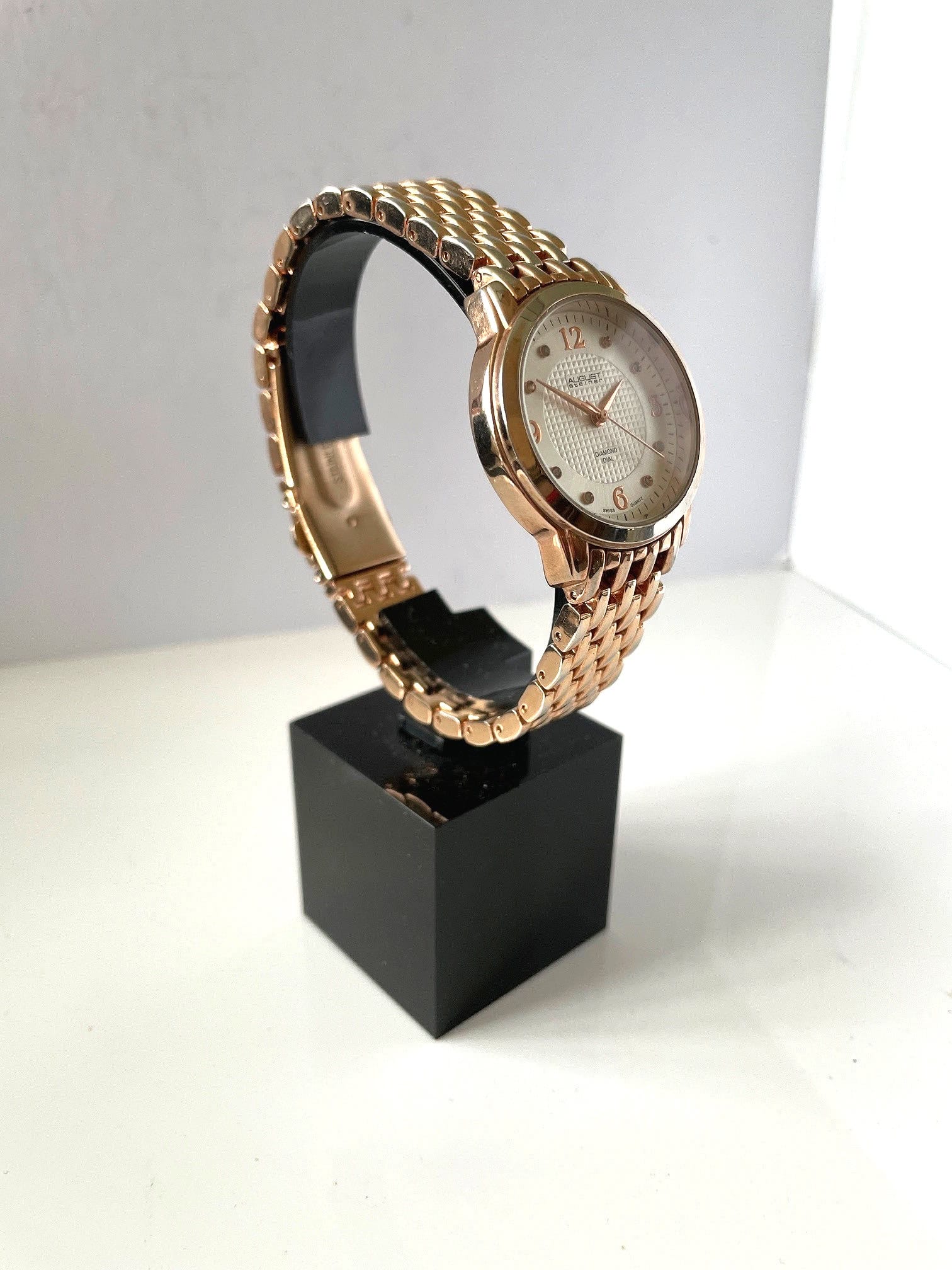 The August Steiner Diamond Silver Dial Ladies' Watch AS8133RG, featuring a gold linked strap and gold hour markers and hands, is displayed on a black rectangular stand against a white background.