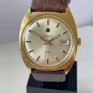 A Roamer Anfibio Mechanical Gold Plated 1960s wristwatch with a cream dial, brown leather strap, and date window at 3 o’clock is displayed upright on a black stand. Buy interest-free in three instalments with Klarna.