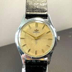 The Marvin Cross Dial Mechanical High-End Watch features a gold-toned face, minimalist hour markers, and a black leather strap, shown upright on a clear stand against a neutral background.