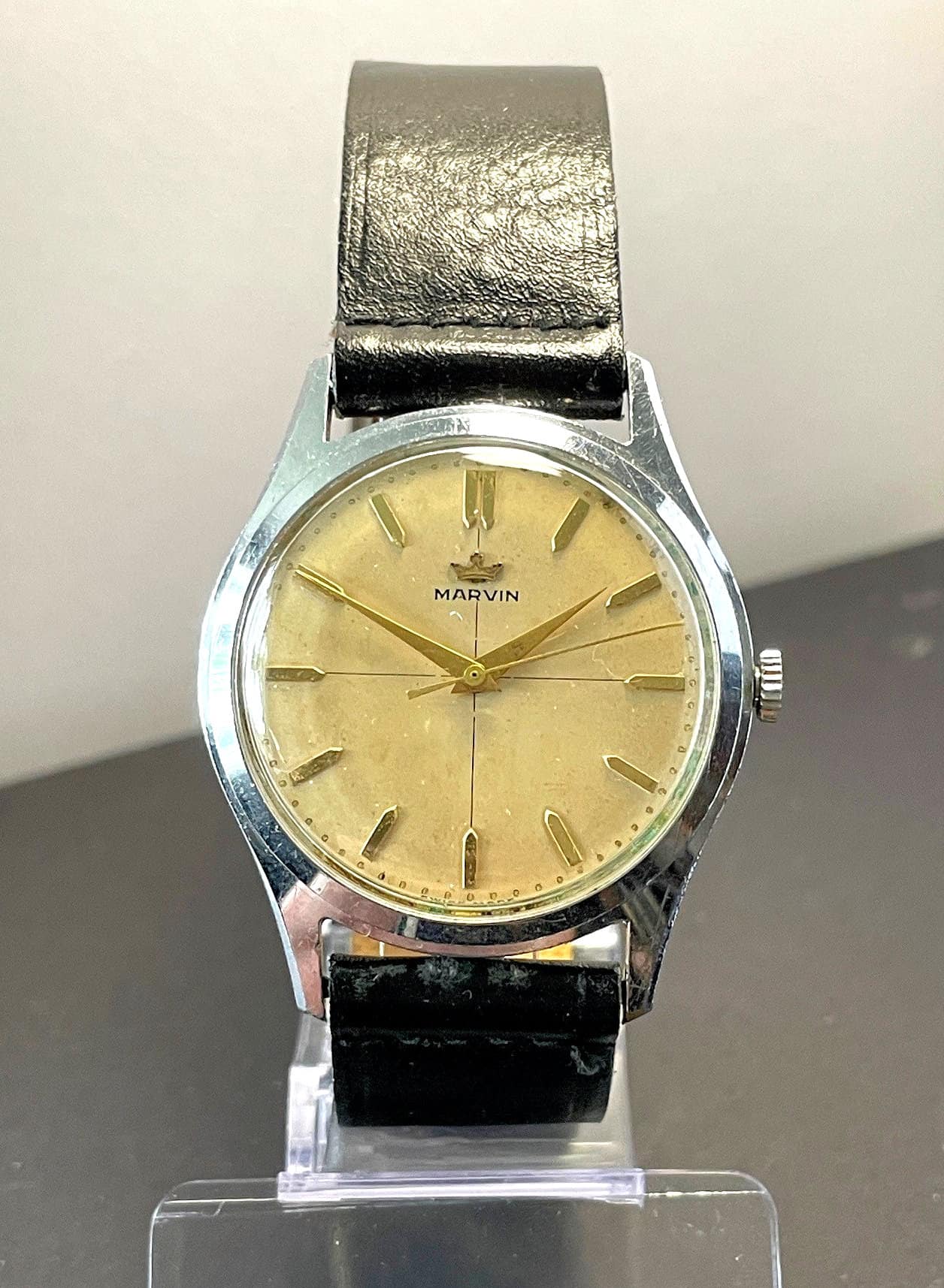 The Marvin Cross Dial Mechanical High-End Watch features a gold-toned face, minimalist hour markers, and a black leather strap, shown upright on a clear stand against a neutral background.