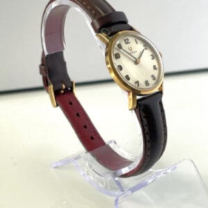 An Omega Ladies 9ct Gold Watch Mechanical c1972, featuring a round gold case and dark brown leather strap, displayed on a clear stand against a plain white background. Pay interest-free in three with Klarna.