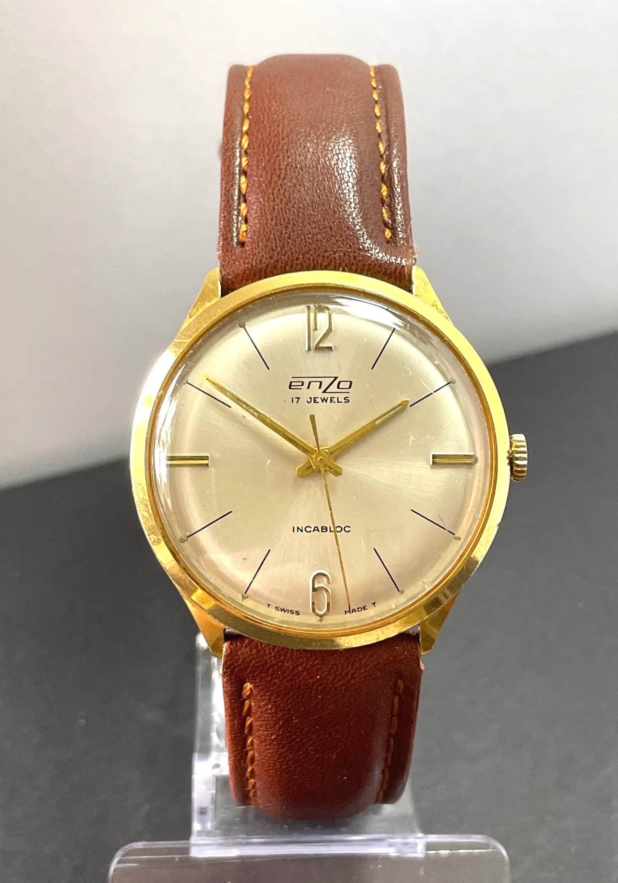 The Enzo 17 Jewel Mechanical Watch features a gold case, light brown leather strap, cream dial with gold markers and thin hands. The watch displays 12:06 and has "17 Jewels" and "Incabloc" on the face.