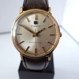 The Tissot Seastar Seven Gold Plated Mechanical features a gold round case, brown leather strap, and silver dial with gold hour markers and hands, displayed upright on a stand. Pay interest-free in three with Klarna.