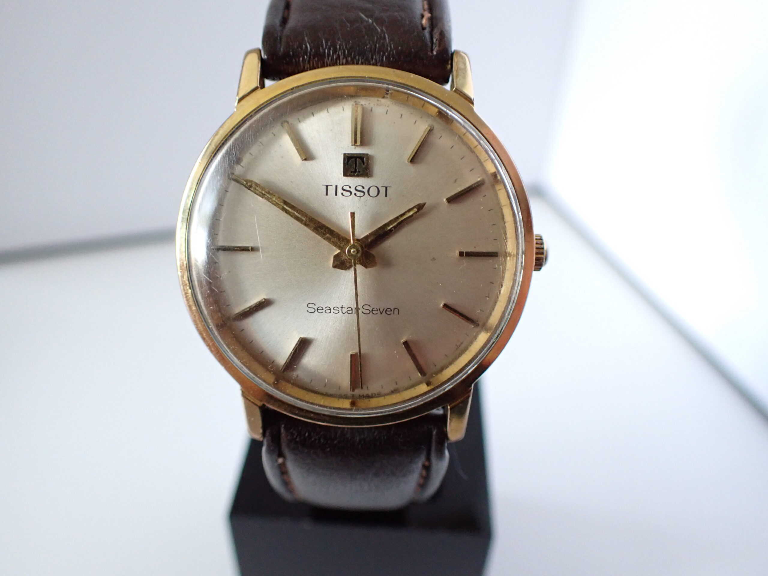 The Tissot Seastar Seven Gold Plated Mechanical features a gold round case, brown leather strap, and silver dial with gold hour markers and hands, displayed upright on a stand. Pay interest-free in three with Klarna.