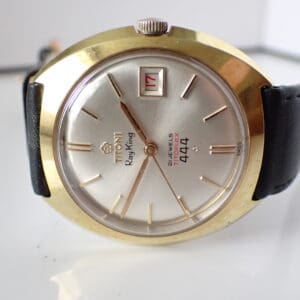 A Titoni Ray King 21 Jewels Titoflex 444 Date wristwatch with a gold-tone case, silver dial, black leather strap, and red “17” date window. Features baton indices, minimalist design. Rare model—pay with Klarna available.