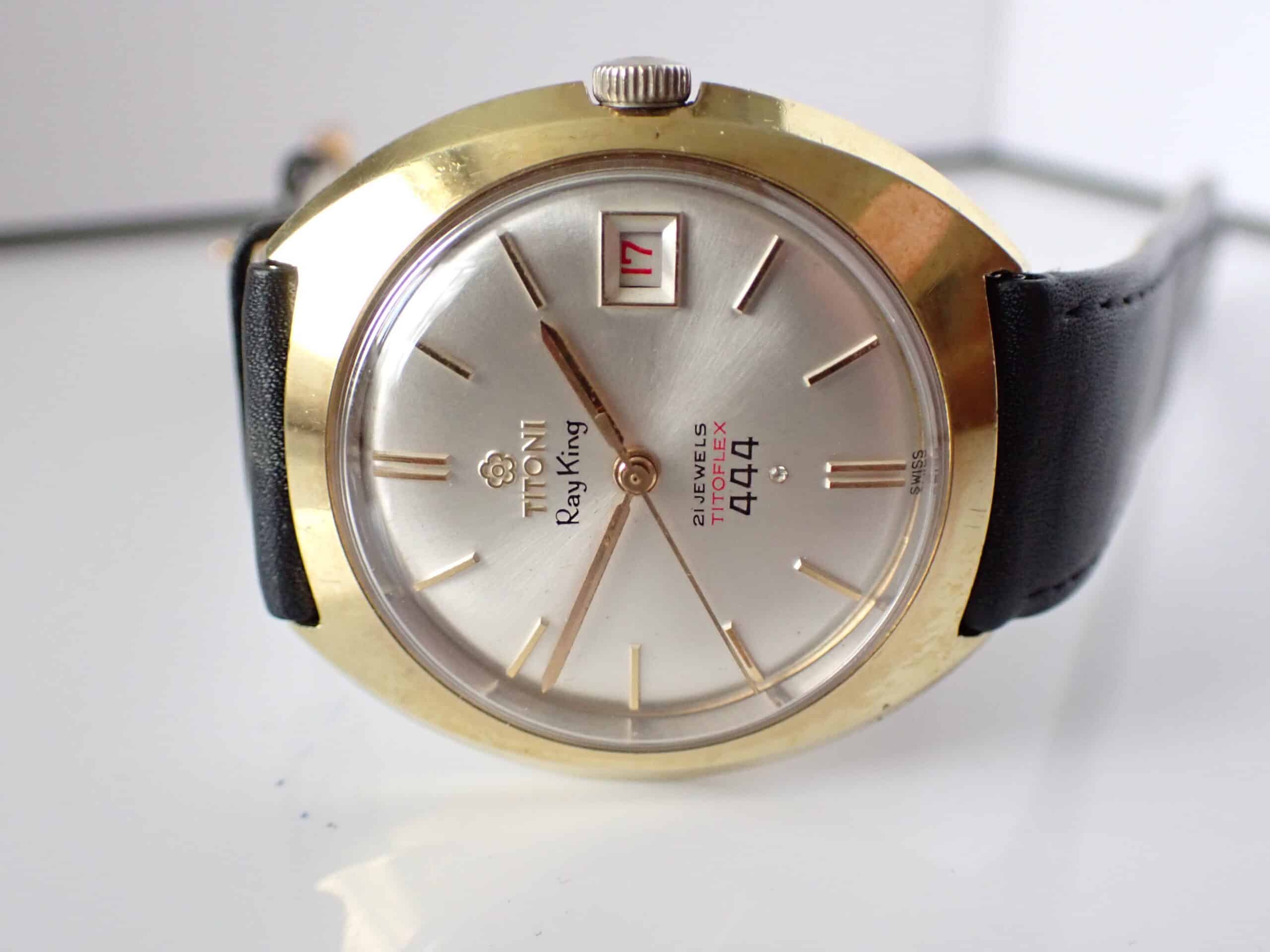 A Titoni Ray King 21 Jewels Titoflex 444 Date wristwatch with a gold-tone case, silver dial, black leather strap, and red “17” date window. Features baton indices, minimalist design. Rare model—pay with Klarna available.
