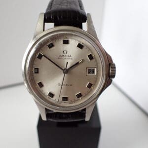 Omega Genève Automatic Watch 166 038 with silver dial, black hour markers, and date window at 3 o’clock on black leather strap, displayed on a stand. Pay with Klarna.