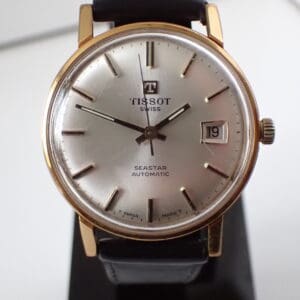 A close-up of the Tissot Seastar Automatic Gold-Plated 1970s Watch, featuring a silver dial, gold-plated case, black leather strap, date display at 19th, and silver hour markers and hands. Available to pay with Klarna.