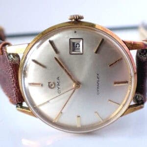 A vintage Cyma Mechanical Cymaflex Date gold-plated watch from the high-end luxury Swiss brand, featuring a round silver dial, gold hour markers, date window at 15, brown leather strap, and "CYMAFLEX" on the face. Pay with Klarna.