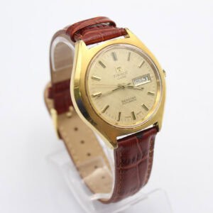 The TISSOT SEASTAR Day Date Automatic features a rare linen beige dial, gold-tone case, and brown leather strap, displayed on a clear stand against a white background. Buy at 0% with Klarna.