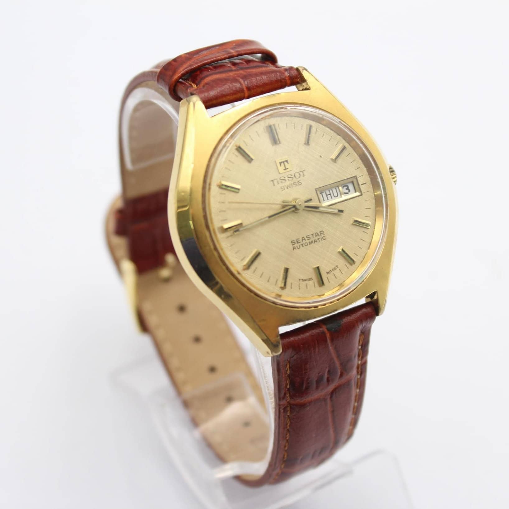 The TISSOT SEASTAR Day Date Automatic features a rare linen beige dial, gold-tone case, and brown leather strap, displayed on a clear stand against a white background. Buy at 0% with Klarna.