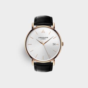 The Prime Ambassador Heritage 1959 is a minimalist wristwatch featuring a white textured dial, gold case, slim gold markers and hands, black leather strap, date display at 3 o’clock, and "Ambassador 1959" printed on the face.