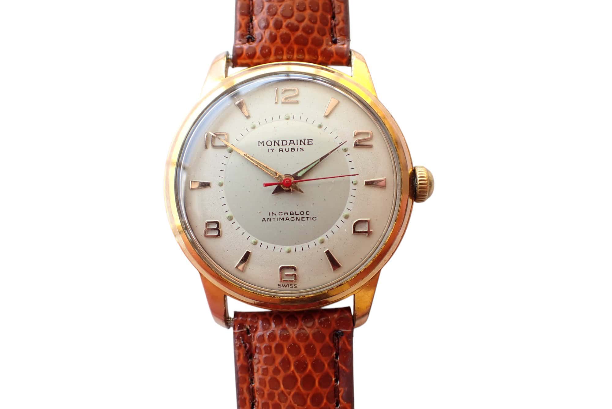 The Mondaine Hand Wind Watch features a brown textured leather strap, gold casing, white dial with gold hour markers and hands, a red second hand, and displays "Mondaine 17 Rubis" on the face.